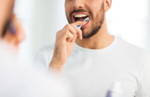 Nose-to-chest view of a man in white t-shirt brushing teeth in front of a mirror